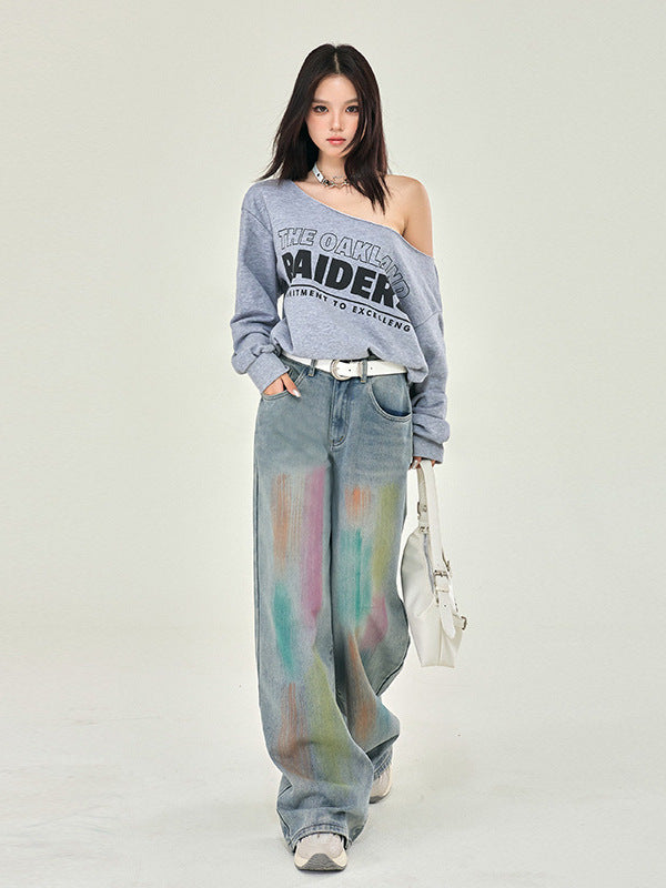 A model poses in oversized Raiders logo sweatshirt, rainbow-washed wide denim pants, white belt, holding white handbag, studio shot