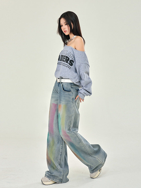 A model in side pose wearing Raiders off-shoulder sweatshirt, watercolor-effect wide-leg jeans, white belt, against neutral backdrop