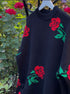 Black sweater with red rose and green leaf pattern hanging against garden backdrop with blooming roses