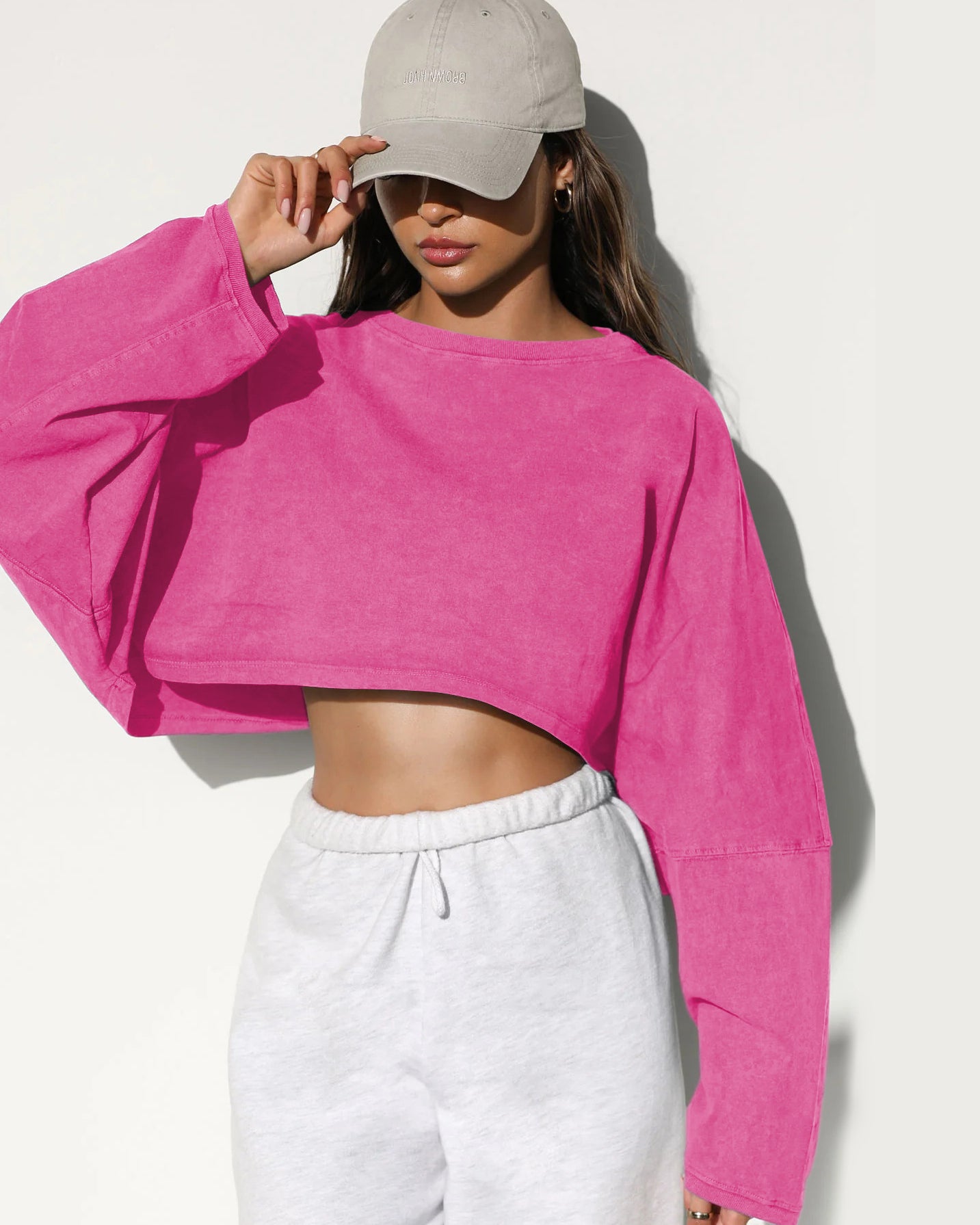 A woman wearing a bright pink cropped long sleeve top and white sweatpants touches her beige baseball cap against a white background