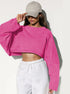 A woman wearing a bright pink cropped long sleeve top and white sweatpants touches her beige baseball cap against a white background