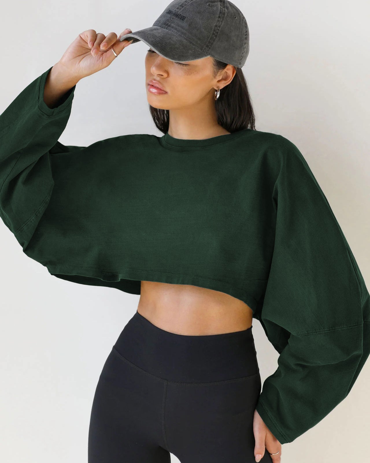 A woman wearing a forest green cropped sweatshirt and black leggings touches her charcoal baseball cap against a neutral background