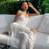 White lace dress with spaghetti straps, worn by a woman sitting on a white couch with green foliage in the background