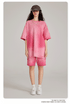 Model wearing pink tie-dye oversized t-shirt with matching shorts, black cap, headphones, gold necklace and white sneakers against neutral background