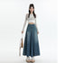 A woman standing with striped pastel cardigan over crop top, blue denim maxi skirt with center pleat detail, platform sandals, and tan shoulder bag