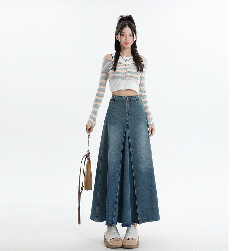 A woman standing with striped pastel cardigan over crop top, blue denim maxi skirt with center pleat detail, platform sandals, and tan shoulder bag