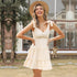 A woman is smiling outdoors in cream mini tiered sundress with tie shoulders, V-neckline and frilled hem, accessorized with straw hat and delicate jewelry
