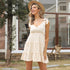 A model is posing against modern architecture backdrop wearing ivory tiered mini dress with shoulder ties, empire waist and ruffle details for summer fashion