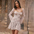 A woman wearing a cream sheer lace mini dress with structured bodice and bell sleeves posing against a rustic stone wall background
