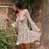 A woman in a cream lace mini dress with flared sleeves examining white roses against a stone wall with climbing vines