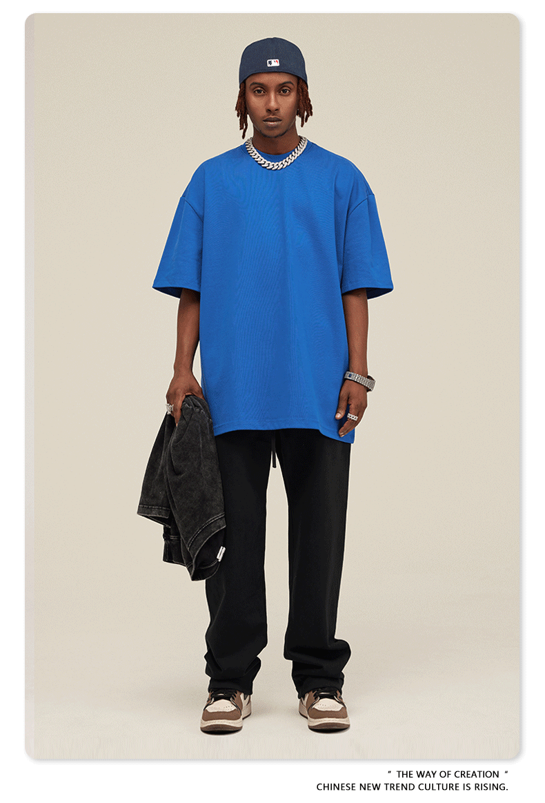 Man with short dreadlocks wearing royal blue oversized t-shirt, black sweatpants, navy baseball cap and silver chain necklace, holding black jacket