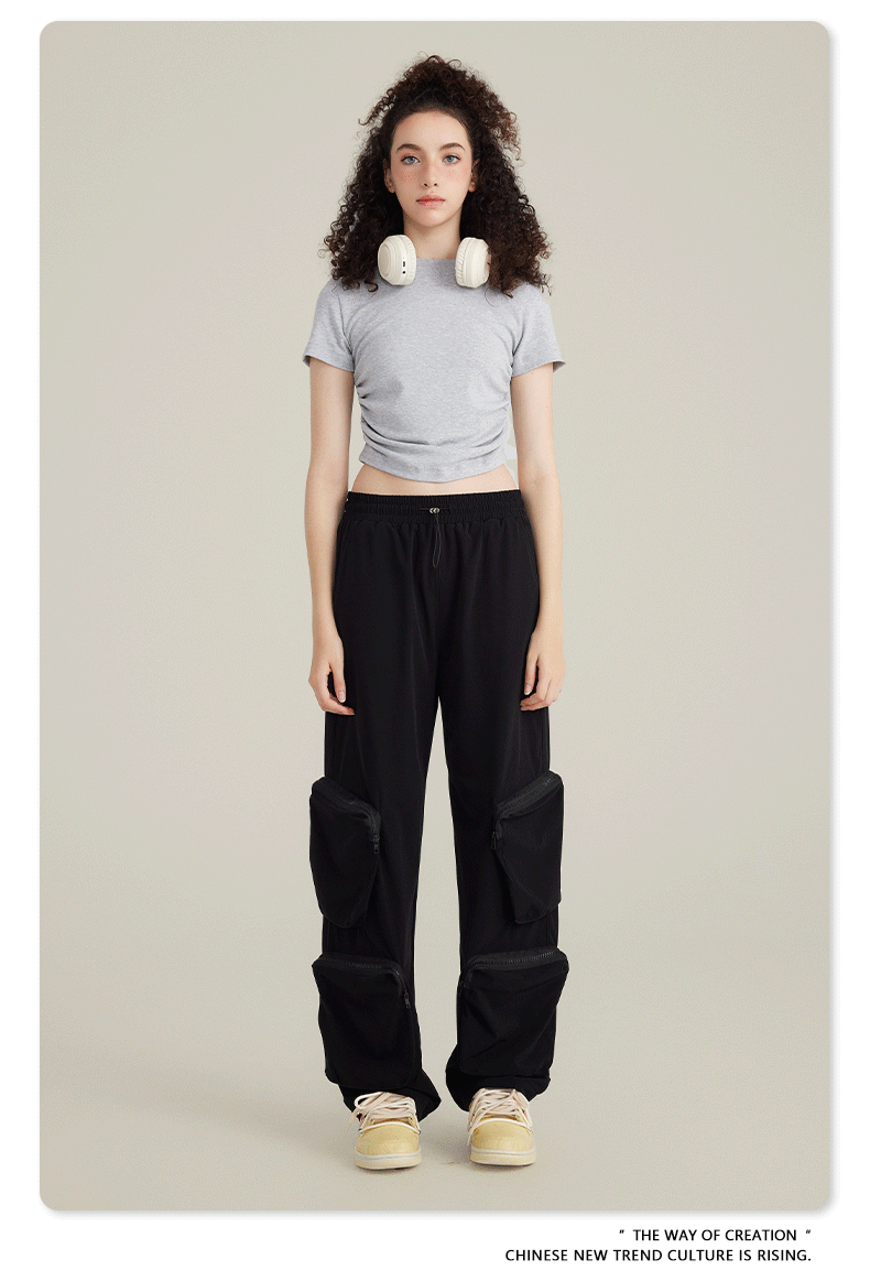 Model with curly dark hair wearing light gray crop top, black cargo pants with zippered pockets, white headphones and cream sneakers on neutral background
