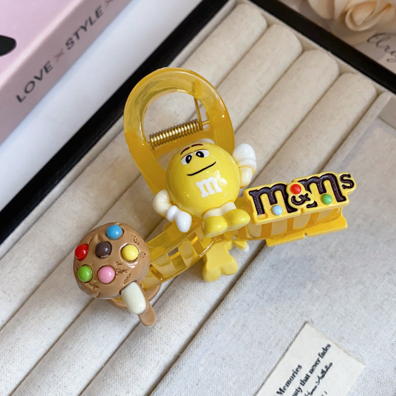 A bright yellow hair claw featuring an M&M character, chocolate chip cookie charm, and candy-themed text.
