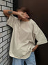 Person wearing a beige oversized shirt and blue jeans against a dark wall.