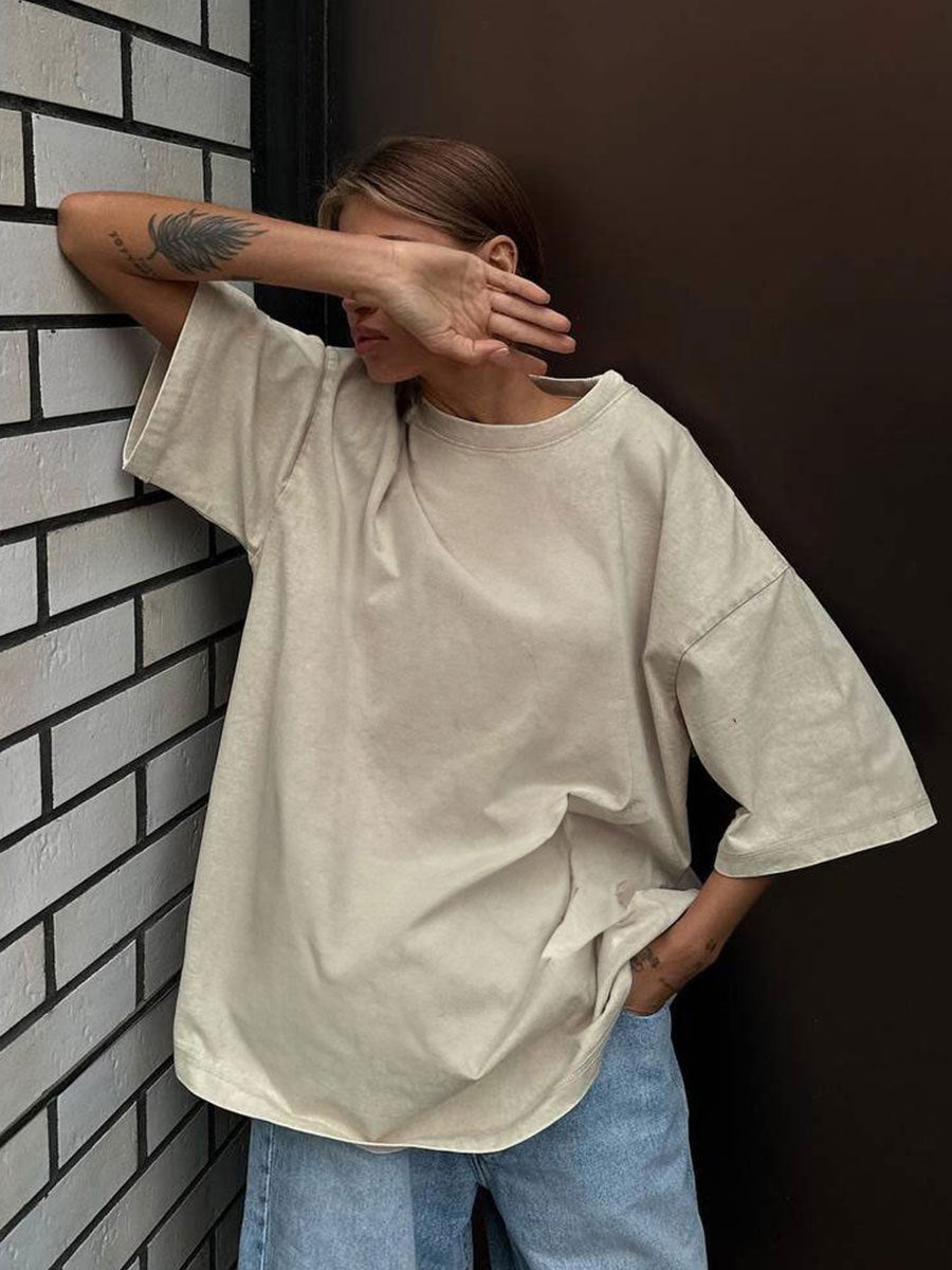 Person wearing a beige oversized shirt and blue jeans against a dark wall.