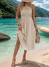 Woman wearing cream halter neck pleated midi dress with straw hat and woven bag, standing on beach with turquoise water views