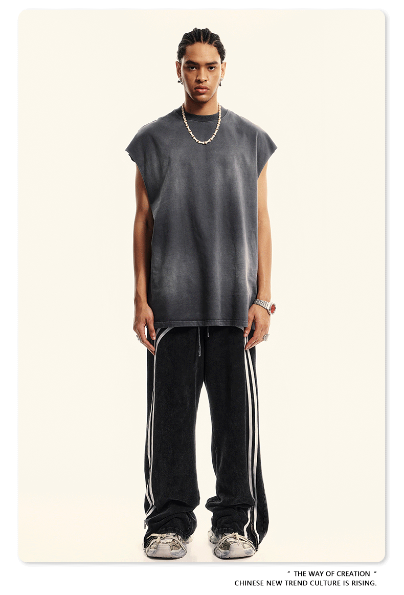 A man wearing a charcoal gray sleeveless muscle tee and black track pants with white stripes stands against a white background