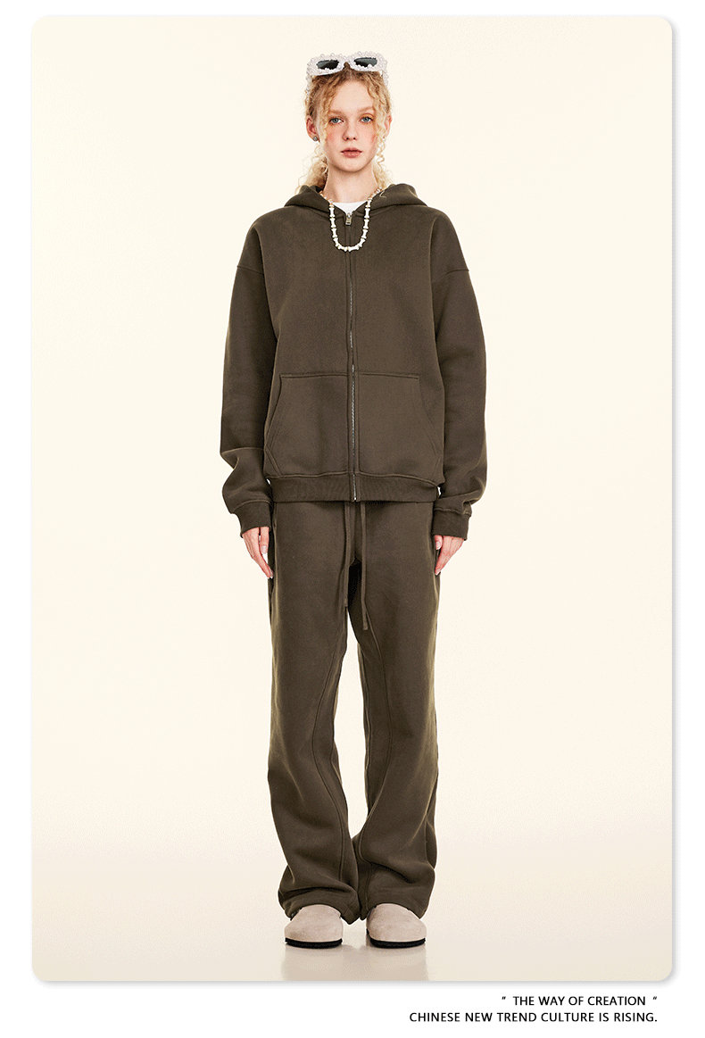 Blonde model wearing olive green oversized zip-up hoodie and matching sweatpants with white sunglasses on head, pearl necklace and beige shoes against cream backdrop