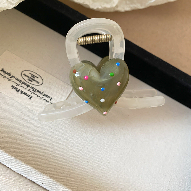 A translucent green hair claw with a 3D heart decorated with pastel polka dots, styled in a model’s brown updo.