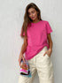Model wears a pink short-sleeve T-shirt paired with high-waisted cream pants, holding a rolled-up magazine against a textured white wall