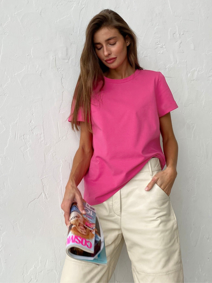 Model wears a pink short-sleeve T-shirt paired with high-waisted cream pants, holding a rolled-up magazine against a textured white wall