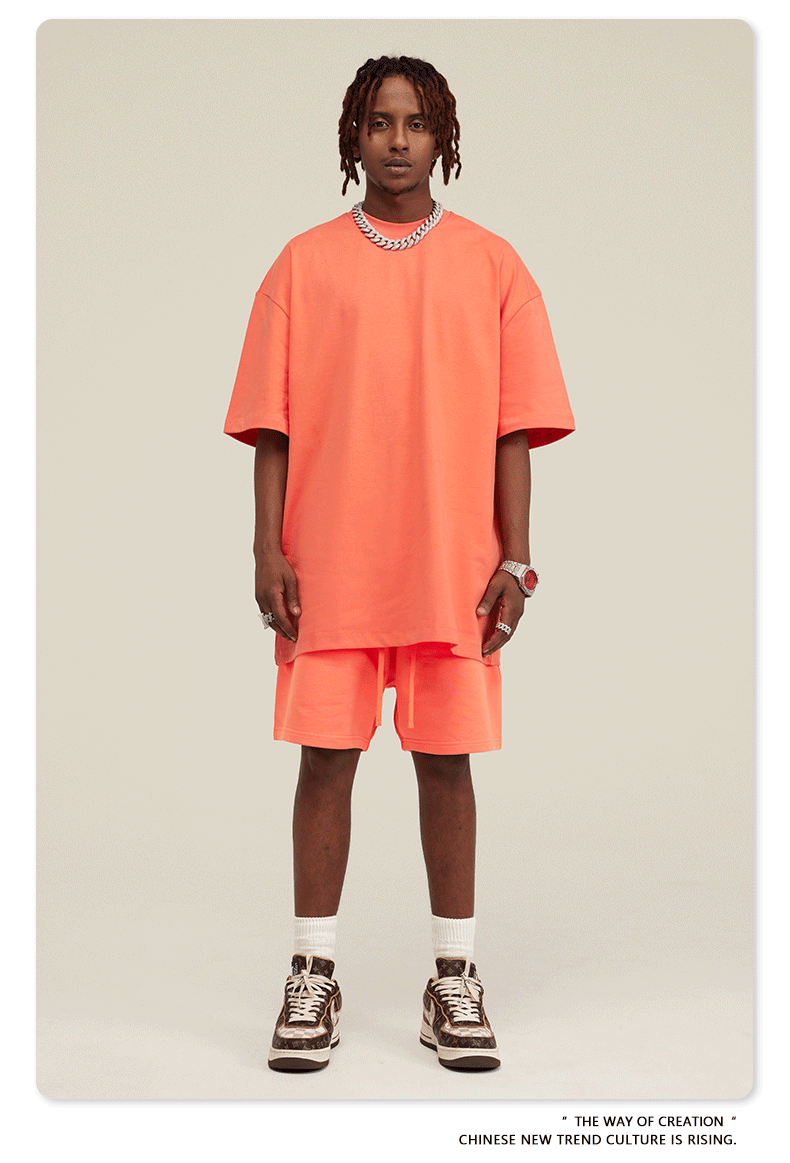 Man with dreadlocks wearing coral/peach oversized T-shirt and shorts set with white sneakers and silver jewelry