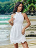 A woman wearing a white eyelet halter sundress with floral embroidery posing on a tropical beach with turquoise water and lush greenery background