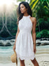 A woman wearing a white eyelet halter mini dress holding a straw hat on a tropical beach with palm trees and rocky shoreline backdrop