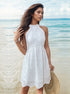 A woman wearing a white eyelet halter dress with scalloped hem standing in shallow ocean water with windswept hair holding a straw hat