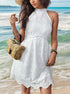 A woman wearing a white eyelet halter dress with scalloped hem holding a woven basket purse while standing in shallow ocean waves
