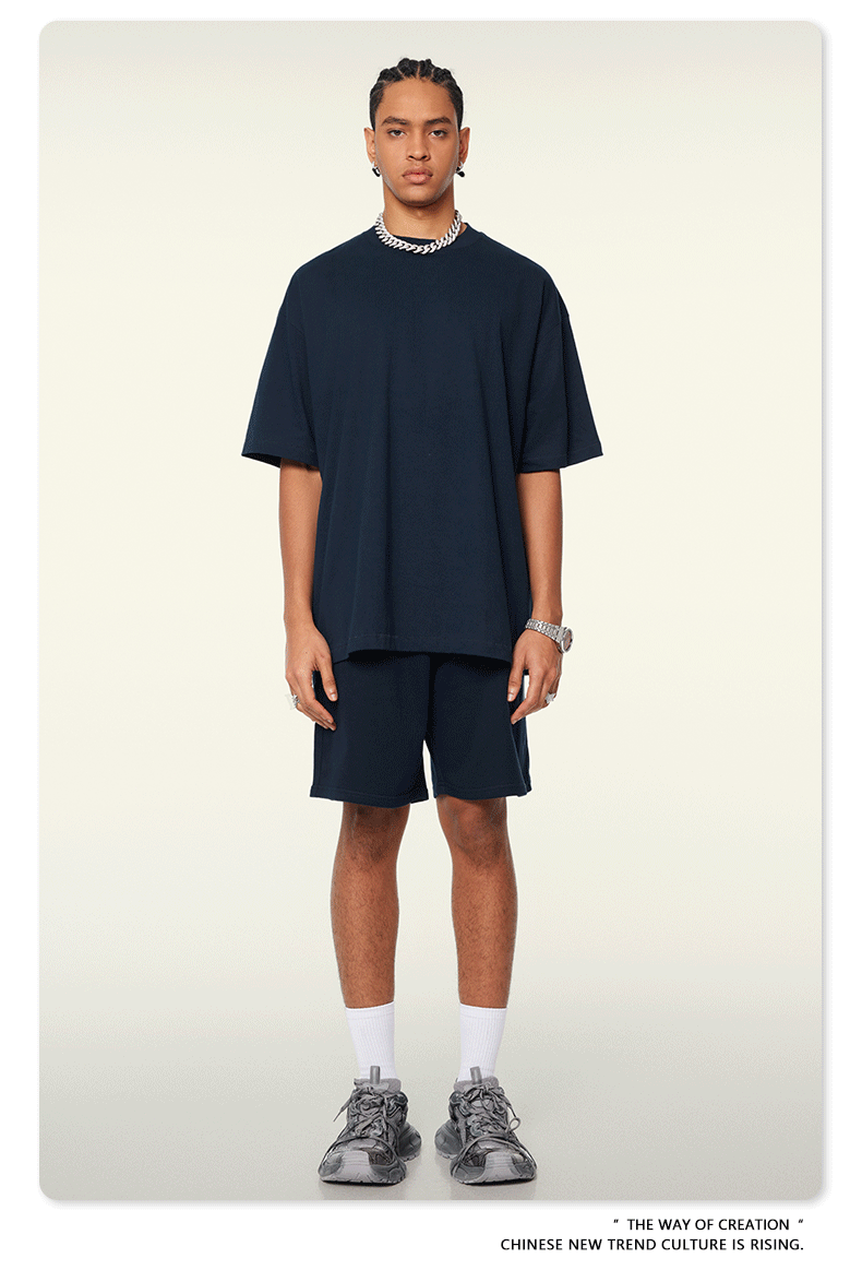 A man with braided hair wearing a navy blue oversized t-shirt and shorts set, accessorized with silver chain, white socks, and athletic sneakers