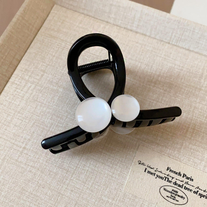 A black hair claw with two white resin beads, placed on a beige surface with a minimalist label.
