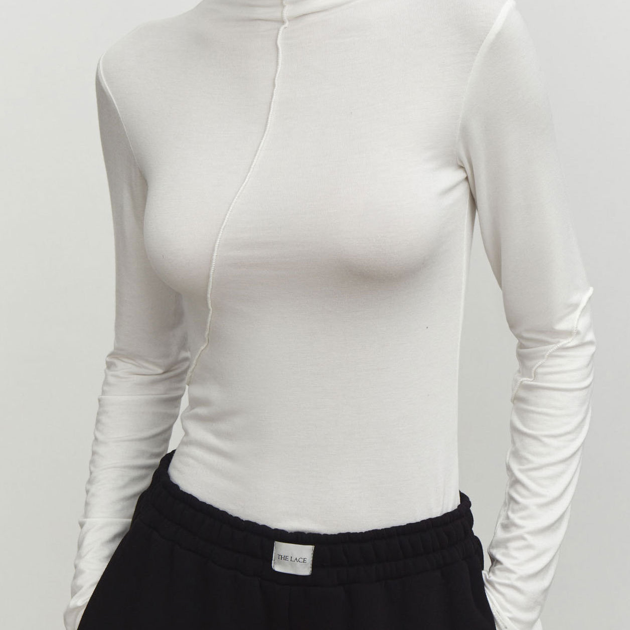 White fitted mock neck top with front seam detail and long sleeves, paired with black pants showing The Lace label