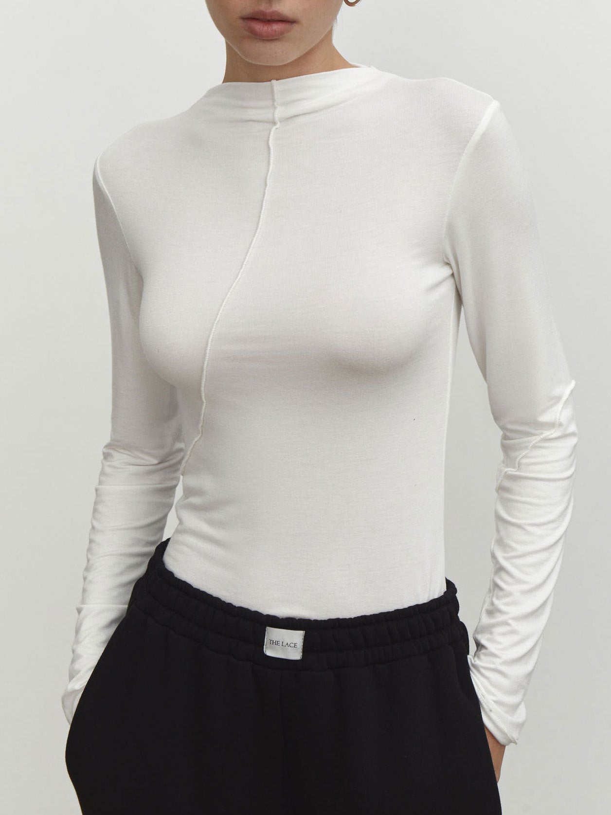 White fitted mock neck top with front seam detail and long sleeves, paired with black pants showing The Lace label