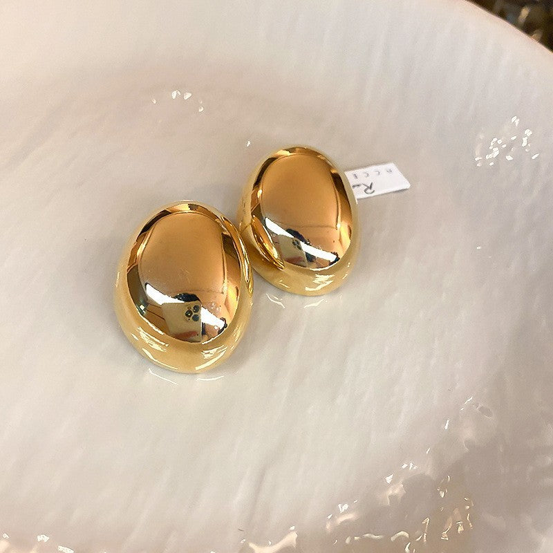Polished Gold Oval Stud Earrings – Simple gold oval stud earrings with a smooth, reflective surface, displayed on a glossy plate
