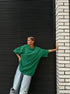 Person wearing a green oversized shirt and light blue jeans leaning against a black shutter.