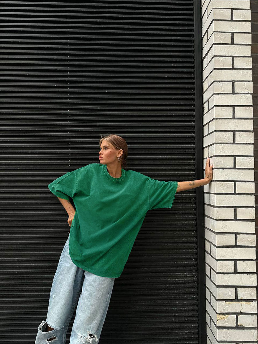 Person wearing a green oversized shirt and light blue jeans leaning against a black shutter.
