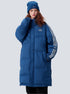 A woman is standing with hand near face against gray backdrop. Wearing royal blue puffer coat with white stripe details, matching beanie and curly red hair