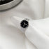 A silver wristwatch-inspired ring with black face displayed on white fabric, featuring detailed band design