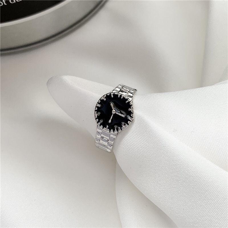 A silver wristwatch-inspired ring with black face displayed on white fabric, featuring detailed band design