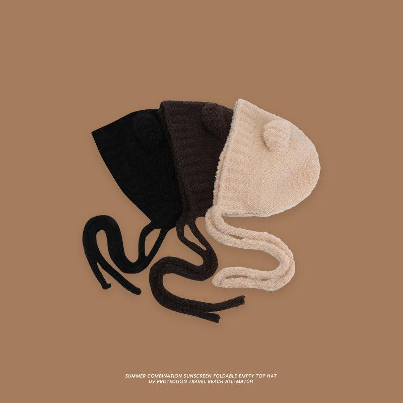 Three plush teddy bear bonnets in black, dark brown, and beige with ear details and chin ties displayed against brown background