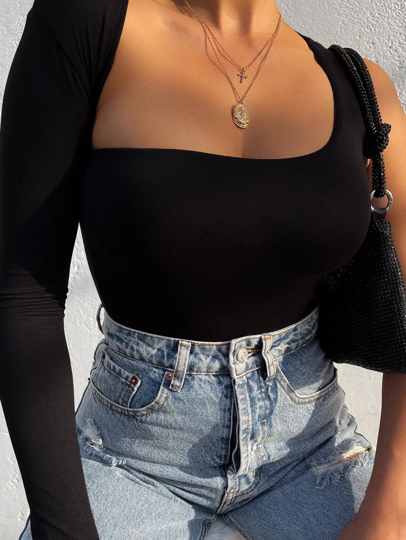 Close-up of a woman in a black asymmetrical bodysuit with long sleeve, accessorized with gold necklaces and a black woven bag.