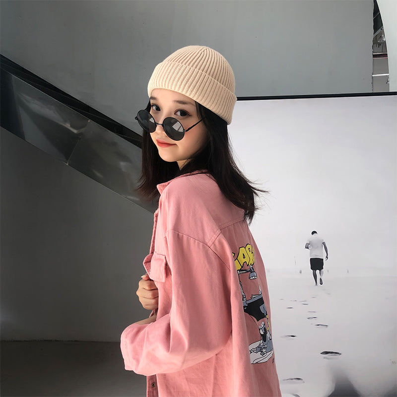 A woman wearing a cream beanie and pink graphic jacket, looking over shoulder with sunglasses, against white studio background