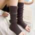 A model is wearing dark gray ribbed leg warmers while sitting on white bedding. One hand visible resting on fabric