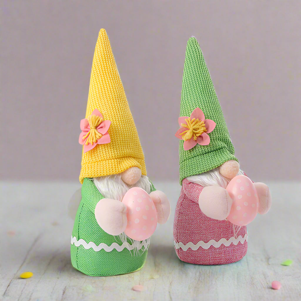 Two Easter gnome plush toys with yellow and green knitted hats, pink flower accents, and holding pink eggs.