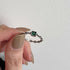 Person holding silver chain link ring with square-cut emerald green stone between fingers against white textured wall