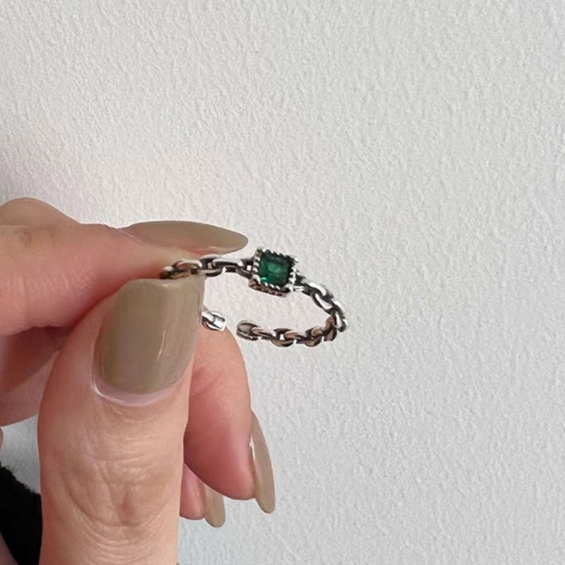 Person holding silver chain link ring with square-cut emerald green stone between fingers against white textured wall