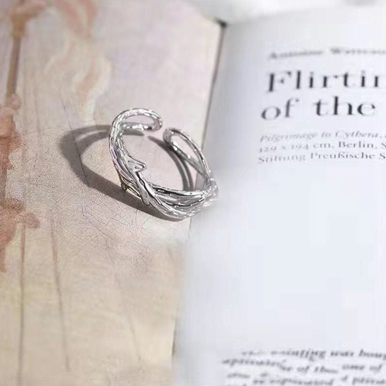 A silver twig-shaped open band ring resting on a book page, with visible text and 'Flirtin' visible in the background
