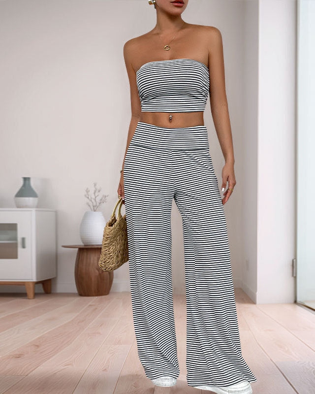 A woman wearing a black and white striped two-piece set, standing in a minimalistic living space with wooden flooring and modern furniture.
