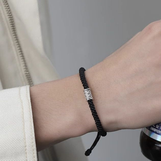 A person wearing a black braided bracelet with a silver charm, dressed in a light-colored jacket.
